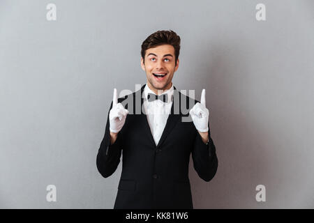 Porträt einer fröhlicher junger Mann im Smoking und Handschuhe, zwei Finger über grauen Hintergrund gekleidet Stockfoto