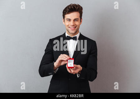 Porträt einer stattlichen glücklicher Mann im Tuxedo holding Open Box mit einem Verlobungsring auf grauen Hintergrund gekleidet Stockfoto