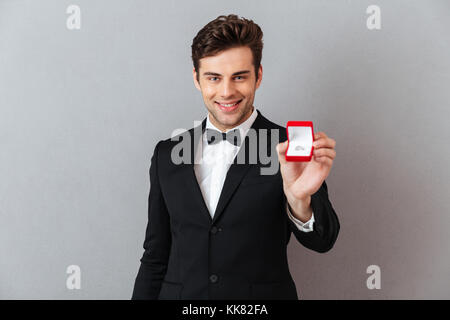 Porträt einer stattlichen glücklicher Mann im Tuxedo holding Open Box mit einem Verlobungsring auf grauen Hintergrund gekleidet Stockfoto