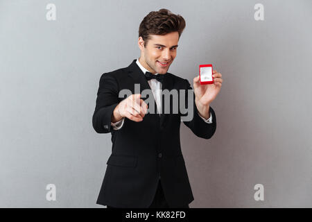 Porträt von einem gutaussehenden charmanten Mann im Smoking holding Open Box mit einem Verlobungsring gekleidet und zeigen mit dem Finger auf Kamera über Grau backgr isoliert Stockfoto