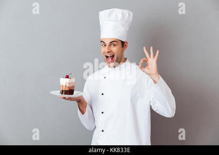 Porträt einer fröhlichen Happy männlichen Chef in Uniform Halteplatte mit Stück Kuchen und zeigen ok Geste über grauen Hintergrund gekleidet Stockfoto