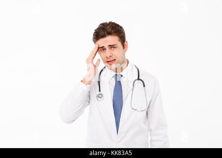 Porträt eines erschöpften jungen männlichen Arzt mit Stethoskop in Uniform leiden unter einer starken Kopfschmerzen gekleidet und Kamera über whi isoliert Stockfoto