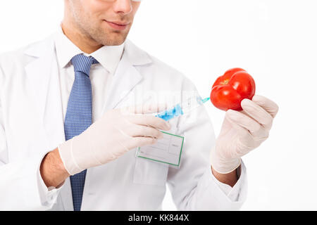 Zugeschnittenes Bild eines lächelnden jungen männlichen Wissenschaftler in Uniform, eine Spritze geschossen in eine Tomate auf weißem Hintergrund gekleidet Stockfoto