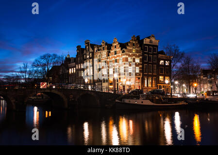 Typische Amsterdamer Gracht Häuser in historischer Architektur entlang der Grachten eine Brücke über das Wasser, in der Blauen Stunde der Dämmerung in Holland beleuchtet. Stockfoto