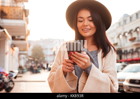 Stilvolle asiatische Frau in schwarzen Hut sms Nachricht auf dem Handy beim Gehen auf die Straße Stockfoto