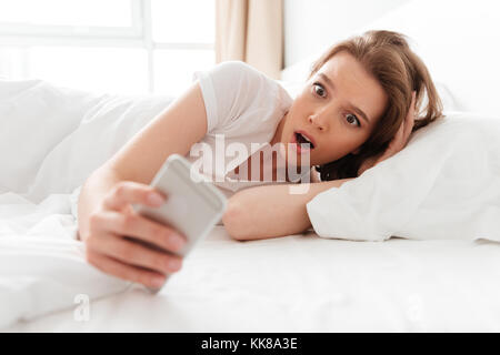 Foto neugierige junge Frau im Bett drinnen zur Seite schauen, Chatten per Handy schockiert. Stockfoto