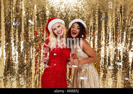 Zwei hübsche junge Mädchen in Weihnachten hüte Toasten mit Champagner Gläser und Kamera über den roten Hintergrund isoliert Stockfoto