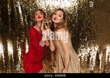 Porträt von zwei freudige hübschen Mädchen in glänzenden Kleidern holding Gläser mit Champagner im Stehen und feiert über golden glänzenden backgrou isoliert Stockfoto