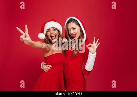 Porträt von zwei aufgeregt verspielten Mädchen in Weihnachten Kleider umarmen und mit Ok Geste über den roten Hintergrund isoliert Stockfoto