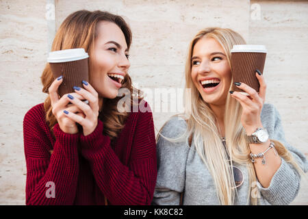 Porträt einer zwei Glückliche lachende Mädchen halten Kaffeetassen auf ihre Gesichter beim Stehen im Freien und bei jedem anderen Suchen Stockfoto