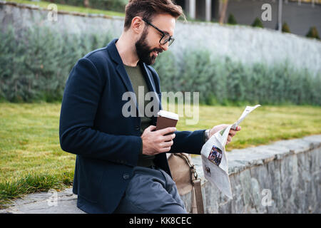 Bild von hübscher junger bärtiger Mann sitzen im Freien Zeitung lesen und Kaffee trinken. Neben der Suche. Stockfoto