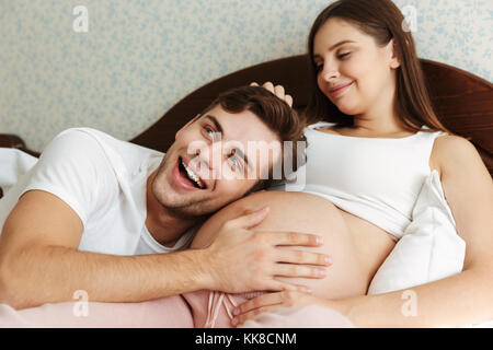 Junge schwangere Frau im Bett lag, mit ihrem Mann im Schlafzimmer zufrieden, Mann liegt auf ihrem Bauch Stockfoto