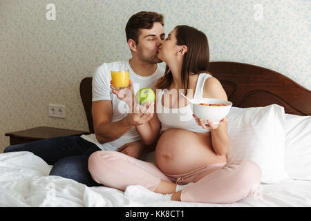 Portrait eines glücklichen jungen schwangeren Paar gesundes Frühstück zusammen, während Sie im Bett sitzen, küssen Stockfoto