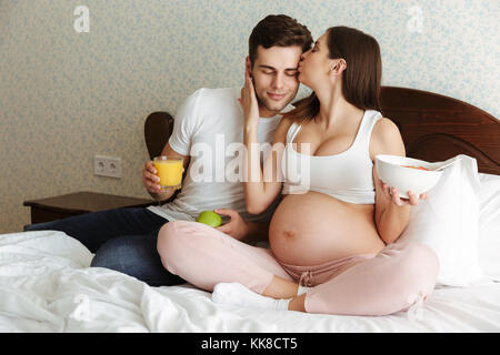 Porträt einer wunderschönen jungen schwangeren Paar gesundes Frühstück zusammen, während Sie im Bett sitzen, küssen Stockfoto