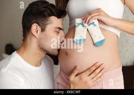 Zugeschnittenes Bild des Menschen hug Bauch seiner schönen schwangere Frau küssen und drinnen zu Hause. Die Augen geschlossen. Stockfoto