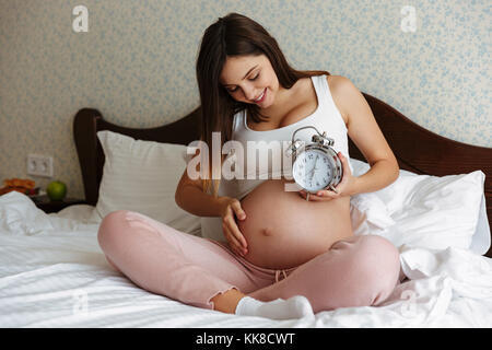 Bild von fröhlichen schwangere Frau zuhause zu Hause sitzen auf dem Bett holding Wecker. Neben der Suche. Stockfoto