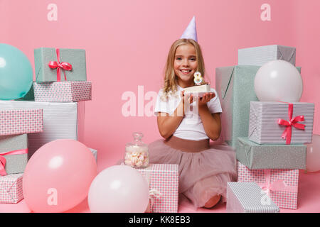 Porträt eines lächelnden hübsches kleines Mädchen in einem geburtstagsmütze Feiern während der Sitzung in einem Stapel von Kisten und Holding Cupcake über rosa isoliert Stockfoto