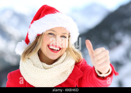 Vorderansicht Porträt einer Frau, die das Tragen von Santa Claus hat an Weihnachten Urlaub mit Daumen hoch mit einem schneebedeckten Berge im Hintergrund Stockfoto