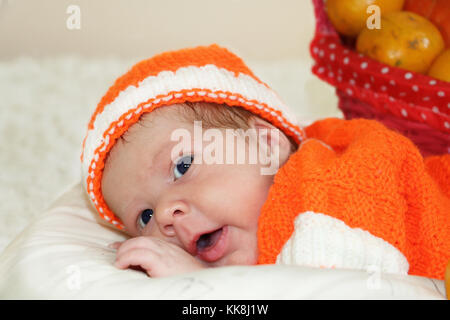 Cute Baby in einen gestrickten orange Kostüm mit Orangen in Korb hinter von ihm angezogen erstaunt. Herbst Halloween oder Ernte Konzept. Baby Gesicht Stockfoto