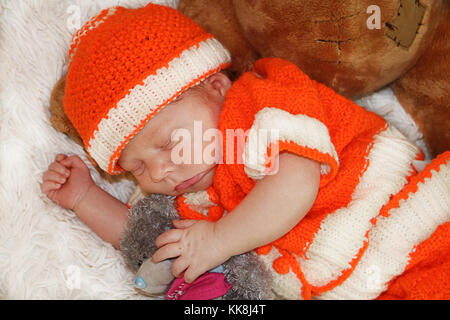 Portrait von niedlichen Neugeborenes im orange Kostüm schlafen auf weißem Fell Decke umarmen Plüschbären. Stockfoto