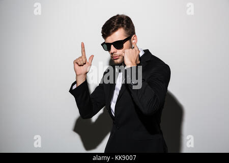 Porträt einer ernster Mann in Ohrhörer und Sonnenbrille mit Gewehr Geste über grauer Hintergrund Stockfoto