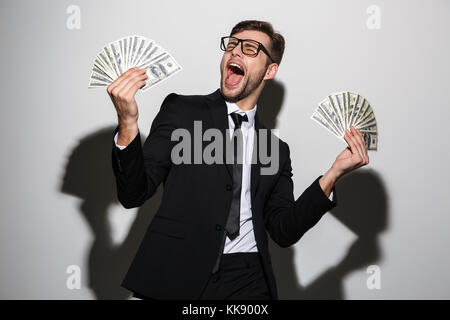 Überglücklich, bärtiger Mann im schwarzen Anzug mit zwei Trauben von Dollar Banknoten, auf weißem Hintergrund Stockfoto