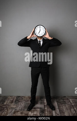 In voller Länge Porträt der Geschäftsmann im schwarzen Anzug, die große Uhr vor seinem Gesicht, über grauer Hintergrund Stockfoto
