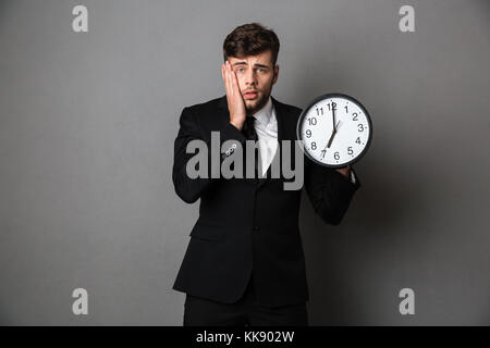 Foto von umgekippt Arbeiter in Anzug holding Uhr schließen, von der Kamera suchen, über grauer Hintergrund Stockfoto