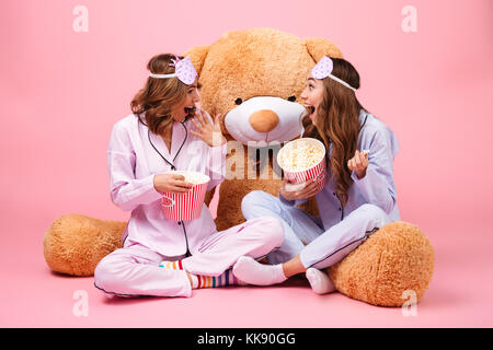 Zwei glückliche hübschen Mädchen im Schlafanzug sitzend mit einem grossen Teddybären und essen Popcorn über rosa Hintergrund gekleidet Stockfoto