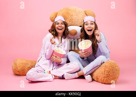 Zwei junge hübsche Mädchen im Schlafanzug lachen während der Sitzung mit einem grossen Teddybären und essen Popcorn über rosa Hintergrund gekleidet Stockfoto