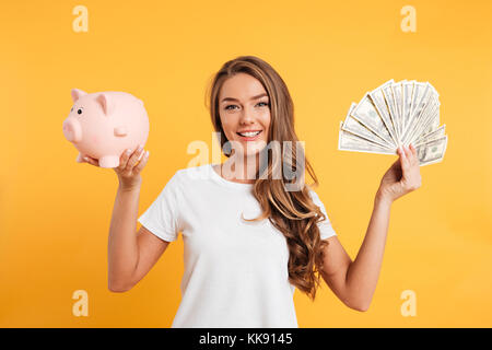 Porträt einer fröhlichen jungen Mädchen, dass Sparschwein und Bündel geld Banknoten isoliert auf gelbem Hintergrund Stockfoto