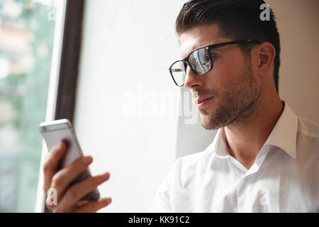 Porträt einer Ernsthafte junge Unternehmer in Brillen mit Handy beim Sitzen im Innenbereich Stockfoto