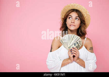 Porträt einer nachdenklich , mädchen im Sommer Hut holding Bündel geld Banknoten und Wegsehen bei copy Space über rosa Hintergrund isoliert Stockfoto