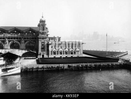 Ein Foto von einem Teil der Fassade des Ellis Island Immigration Prüfplatz, der französischen Renaissance Revival Gebäude mit roten Ziegeln und Kalkstein Verkleidung konstruiert wird, das Gebäude wurde erstmals am 17. Dezember 1900 eröffnet, ist im Jahr 1954 war nach der Verarbeitung über 12 Millionen Einwanderer die Einreise in die Vereinigten Staaten geschlossen, Türme an den Ecken des zentralen Teil der Struktur mit einem überdachten Gang führt zum Haupteingang gesehen werden können, ein großes Boot ist am Ende des Laufstegs angedockt ist, eine grosse Gruppe von Leuten gesehen, wartet das Boot, New York, 1907 beenden zu können. Von Stockfoto