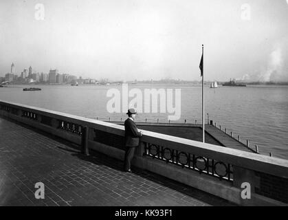 Schwarz-weiß Foto mit einem Mann in Anzug und Derby hat unter Beachtung der Hafen aus der Beobachtung Dach der Einwanderung, die Skyline von New York, in dem die fast abgeschlossenen Woolworth Building Tower, auf der linken Seite zu sehen ist, von Edwin Levick, Ellis Island, New York, 1907. Von der New York Public Library. Stockfoto