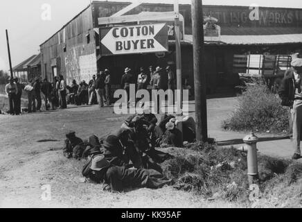 Schwarz-weiß Fotografie afroamerikanische Männer stehen auf einer Schotterstraße, vor einem großen, 1-stöckiges Gebäude, ein Schild mit der Aufschrift 'Baumwolle Käufer' im Vordergrund, 1939. Von der New York Public Library. Stockfoto