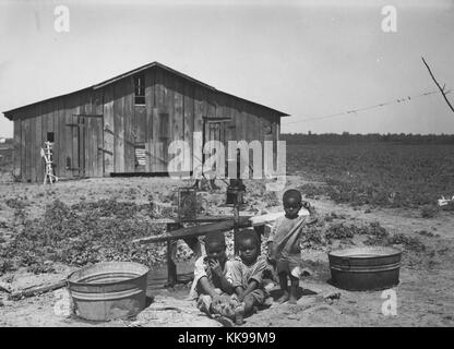 Schwarz-weiß Foto von drei Afrikaner - junge Kinder, zwei Sitzen, einen ständigen, im Freien, eine Wasserpumpe und hölzerne Struktur können im Hintergrund gesehen werden, West Memphis, Arkansas, 1935. Von der New York Public Library. Stockfoto