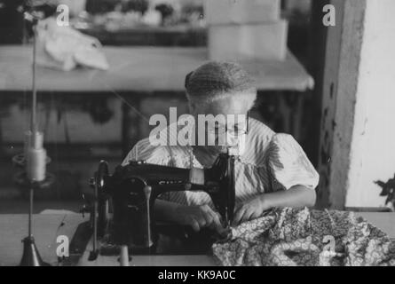 Ein Foto von einem ausgereiften Puerto Rican Frau mit einer Nähmaschine in die Everglades Handarbeit Fabrik, sie ist mit der Maschine ein Stück Blumen gemusterte Stoff zu arbeiten, trägt sie eine gestreifte Bluse und Gläser, ein weiteres Werkbank in der Fabrik hinter ihr gesehen werden kann, San Juan, Puerto Rico, 1864. Von der New York Public Library. Stockfoto