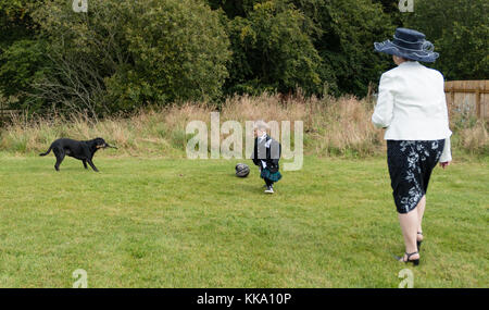 Kleine Jungen in einem Kilt Fußball spielen mit einem Hund, von einer Dame in einen Hut beobachtete Stockfoto