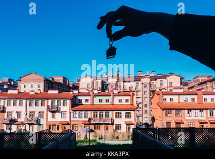 Männliche Hand, die Hausschlüssel auf Haus geformte Schlüsselbund. Menschliche Hand silhouette Holding Schlüsselbund in Form von Haus mit Schlüssel auf dem Hintergrund ein neues Gebäude Stockfoto