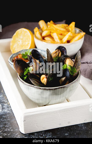 Miesmuscheln mit Kräutern in eine Schüssel geben und mit Zitrone und Pommes frites auf ein weißes Holzbrett. Meeresfrüchte essen am Ufer des französischen Meer. dunklen Hintergrund Stockfoto