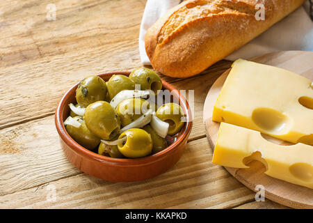 Großen spanischen Grünen gordal Oliven mit Kräutern und Zwiebeln in Steingut Schale baguette maasdamer Käse auf Schneidebrett Holz Tisch rustikale Einrichtung mediterr Stockfoto