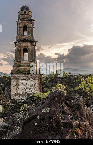 Der Turm der Kirche San Juan Parangaricutiro, der bei Sonnenuntergang aus einem Meer aus getrocknetem Lavagestein im abgelegenen Dorf San Juan Parangaricutiro, Michoacan, Mexiko, aufsticht. Diese Kirche ist die einzige noch verbliebene Struktur, die beim achtjährigen Ausbruch des Vulkans Paricutin begraben wurde, der 1943 zwei Dörfer verzehrte und die Region mit Lava und Asche bedeckte. Stockfoto