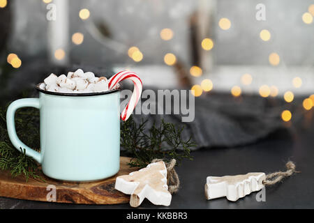 Emaille Tasse heißen Kakao trinken mit Marshmallows und Candy Cane gegen eine rustikale Hintergrund mit schönen Bokeh Lichter vor einem Fenster. Holz chri Stockfoto