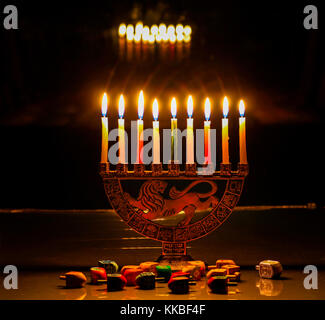 Hanukkah Menorah mit brennenden Kerzen auf dem Tisch sitzt; die Lichter spiegeln sich in den dunklen Fenster über ihnen; dreidels auf dem Tisch vor o verstreut Stockfoto