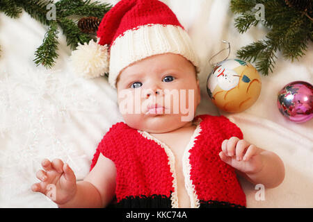 Baby erstes Weihnachten. Schönes kleines Baby in Santa Hut feiert Weihnachten. Das neue Jahr Urlaub. Baby mit Weihnachtsmütze im Inneren Lametta, Kugeln und und Stockfoto