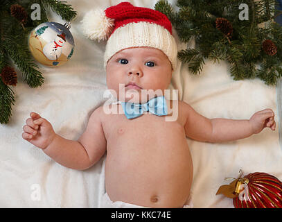 Baby erstes Weihnachten. Schönes kleines Baby in Santa Hut und Fliege legt auf weißem Hintergrund mit Weihnachtsbaum Nadeln und Kugeln eingerahmt. Das neue Jahr Stockfoto