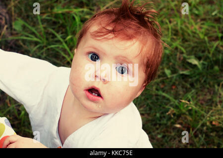 Portrait von Red überrascht vorangegangen Baby mit großen blauen Augen und geöffnetem Mund in weißem Hemd bekleidet, auf natürlichen, grünen Hintergrund mit Gras. Stockfoto