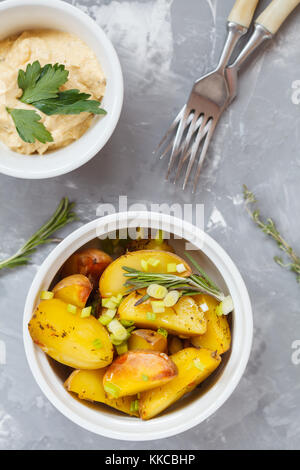 Gebackene Kartoffeln mit Rosmarin und grüne Zwiebeln aus weisser Keramik Form mit Hummus, Ansicht von oben, grauen Hintergrund. gesund vegan detox food Konzept. Stockfoto