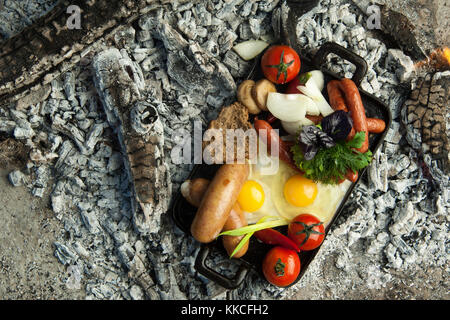 Geräucherte Würstchen mit Tomaten und Eiern liegen auf Holzkohle. Das Gericht wird gekocht und geräuchertem auf Holzkohle Stockfoto